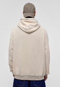 Beige oversized hoodie met een grote capuchon en geribbelde manchetten, gecombineerd met een grijze baseballpet. Zachte textuur, casual ontwerp en zichtbare naden.