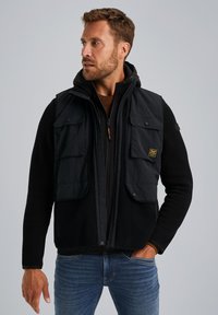 Chaleco negro sin mangas con múltiples bolsillos frontales, superpuesto a una chaqueta con capucha negra. Combinado con jeans azules y un suéter marrón texturizado.