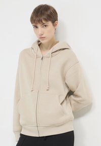 Felpa con zip beige, dotata di cappuccio con cordino, tasche frontali e polsini a coste. Texture morbida e vestibilità rilassata, adatta per un abbigliamento informale.
