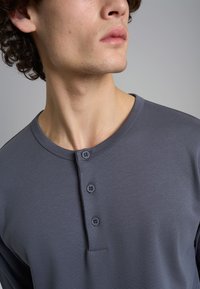 Maglietta grigia a maniche lunghe con scollatura tondeggiante, abbottonatura a tre bottoni, texture liscia e design minimalista.