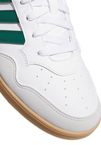 Witte leren sneaker met groene strepen, witte veters, geperforeerde neus en lichtbruine rubberen zool, met een close-up detail.