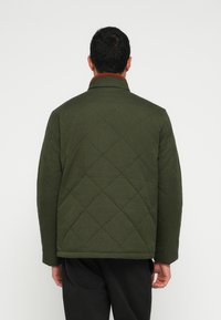 Casaco acolchoado verde-oliva com gola alta e tecido texturizado. Apresenta um padrão de diamante e um detalhe castanho no pescoço.