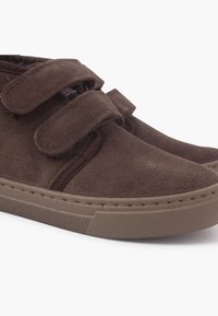 Zapatos de ante marrón con dos tiras de velcro, punta redondeada y suela de goma texturizada. Presentan costuras sutiles a lo largo de los bordes.