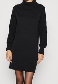 Robe en maille noire à col roulé avec manches texturées, poignets et ourlet côtelés. Longueur au-dessus du genou, avec une coupe décontractée et une couleur unie.