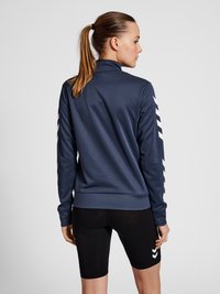 Marinblå atletisk jacka med hög krage och vita chevronränder på ärmarna, kombinerad med svarta shorts. Slät textur, figurnära design.