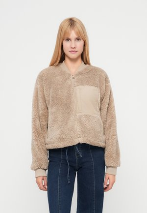 Beige, fuzzy bomber jacket with a zip front, rounded collar, and a solid beige chest pocket. Ribbed cuffs and bottom hem. Soft texture.
