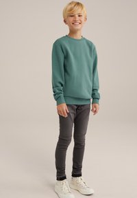 Sweatshirt vert à col rond, avec des poignets et un ourlet côtelés ; associé à un pantalon gris ajusté et des baskets montantes blanches. Tenue simple et décontractée.