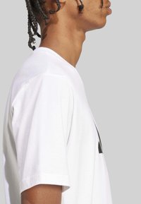 Camiseta de algodón blanca con cuello redondo, mangas cortas y un elemento gráfico negro en la parte delantera. Textura suave.