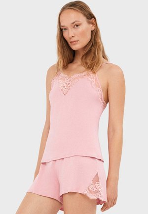 Mujer vestida con camisolín rosa con encaje y pantalones cortos a juego, de pie contra un fondo claro y liso.