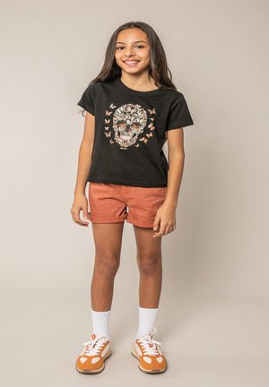 Meisje staat en lacht, draagt een zwart T-shirt met een ontwerp van een bloemen schedel en vlinders, roestkleurige korte broek, witte sokken en oranje sneakers.