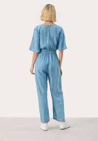 Mono de mezclilla azul claro con mangas cortas, cintura ceñida y cierre de botón en la parte posterior. Combinado con zapatillas blancas.