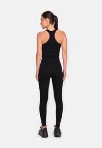 Musta urheilullinen tankkitoppi, jossa on urheilullinen selkädesign, yhdistettynä korkeavyötäröisiin mustiin leggareihin. Mallilla on päällään mustat kengät, joissa on teksturoidut pohjat.