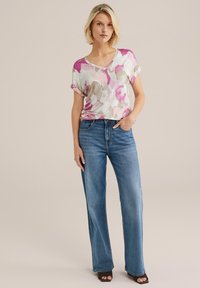 Majica kratkih rukava s V-izrezom od lagane tkanine s ružičastim i bež cvjetnim uzorkom, uparena s opuštenim plavim trapericama i crnim sandalama otvorenih prstiju.