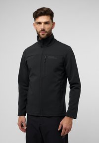 Schwarze Softshell-Jacke mit hohem Kragen, Reißverschluss vorne und Logo auf der Brust. Hat eine taillierte Passform und eine glatte Textur.