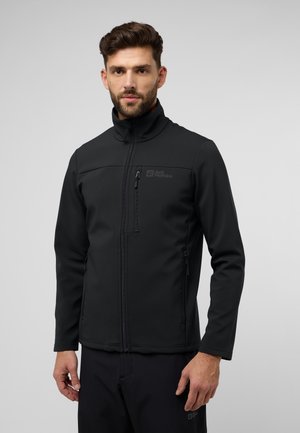 Veste softshell noire avec un col haut, fermeture éclair à l'avant et logo sur la poitrine. Présente une coupe ajustée et une texture lisse.