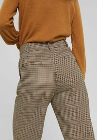 Rutmönstrade byxor i brun, krämfärgad och svart, med hög midja, bakfickor och skräddarsydd passform. Texturerad tyg med ett klassiskt mönster.