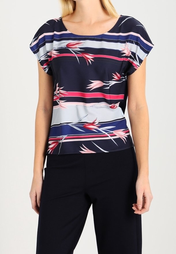Femme portant un chemisier bleu marine avec des rayures florales roses et rouges, et un pantalon noir, debout devant un fond clair uni.