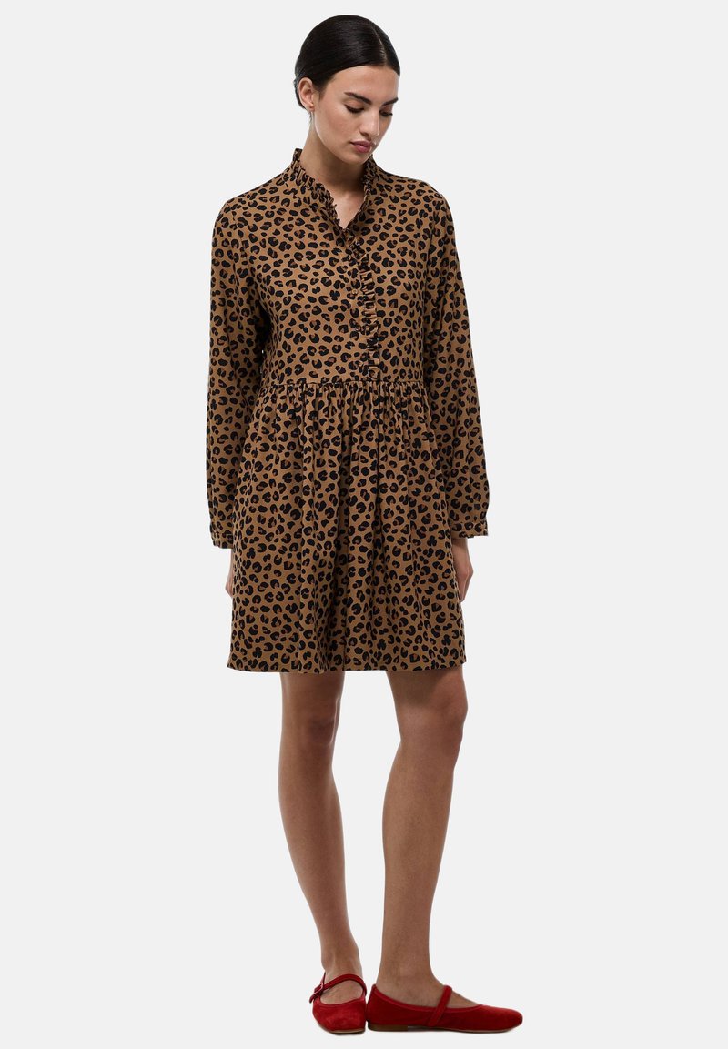 Robe imprimée léopard en brun et noir, dotée d'un col, d'un système de boutonnage sur le devant, de manches longues et d'une taille froncée. Associée à des ballerines rouges.