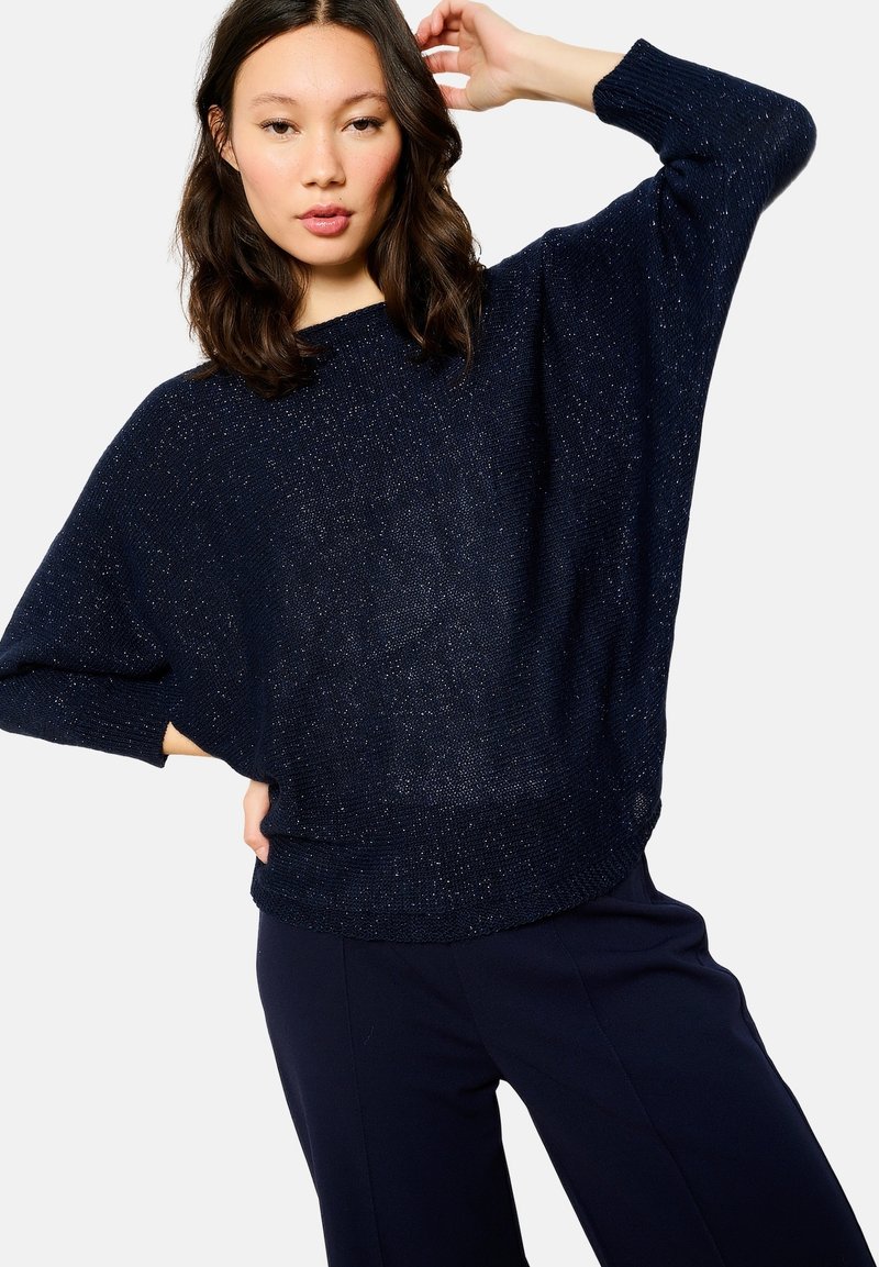 Navy blue oversized knit sweater with a textured finish, wide sleeves, and a relaxed fit. Paired with navy trousers for a cohesive look.