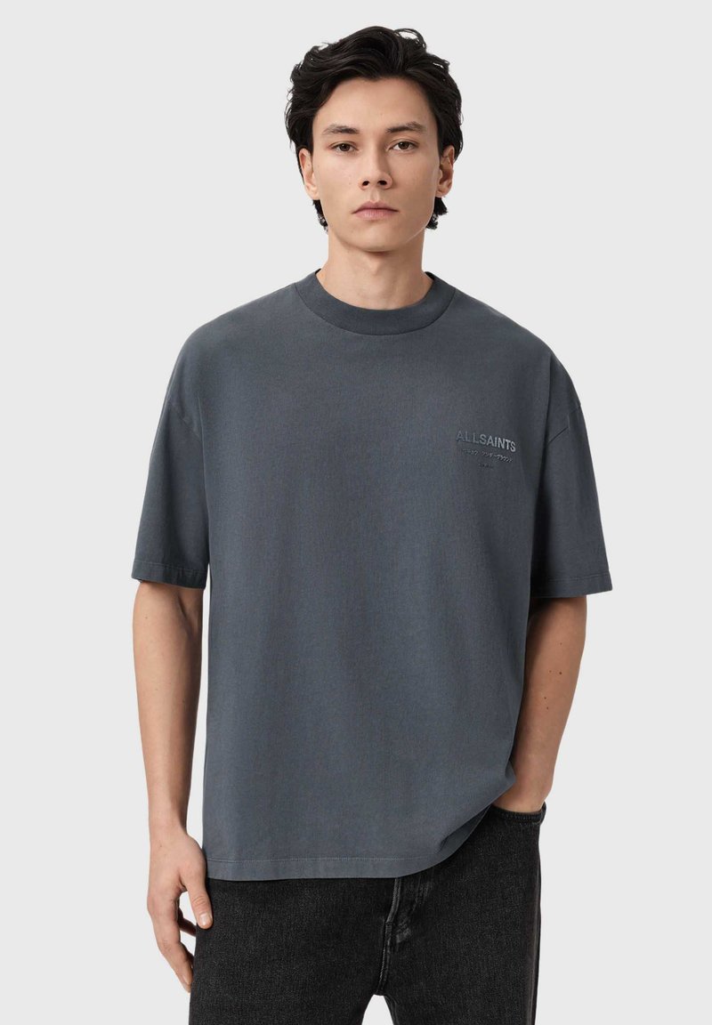 Junger Mann mit dunklen Haaren trägt ein übergroßes dunkelgraues T-Shirt mit dezentem Text auf der Brust und schwarze Jeans, steht vor einfachem Hintergrund.