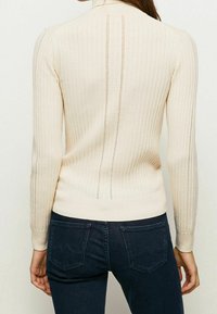 Pull-over crème côtelé avec manches longues et col montant. Présente un motif ajouré vertical à l'arrière et une silhouette ajustée.