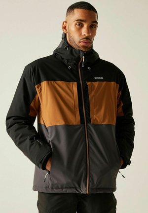 Mann trägt eine schwarz-braun-grau farblich blockierte Kapuzenjacke mit Händen in den Taschen, schaut nach vorne vor einfarbigem Hintergrund.