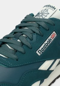 Tirkizne Reebok Classic tenisice s teksturiranim vezicama, bijelim potplatom i logom na bočnoj strani koji prikazuje zastavu Ujedinjenog Kraljevstva.