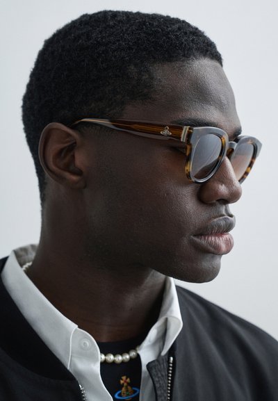 Ochelari de soare cu ramă dreptunghiulară în model tartaruga, având un accent argintiu pe braț. Purtați cu o jachetă bomber neagră și cămăși suprapuse.