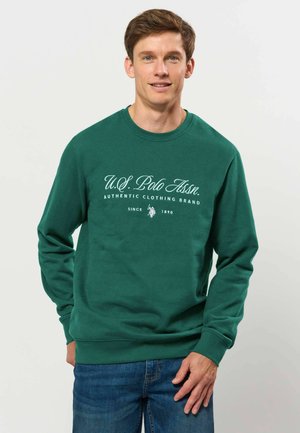 Jeune homme portant un sweat-shirt vert U.S. Polo Assn. et un jean bleu, debout devant un fond clair uni.
