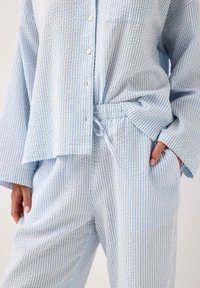 Ljusblå och vitrandig pyjamasuppsättning med knäppskjorta, långärmade ärmar och byxor med elastiskt midjeband, tillverkad av mjukt strukturerat tyg.
