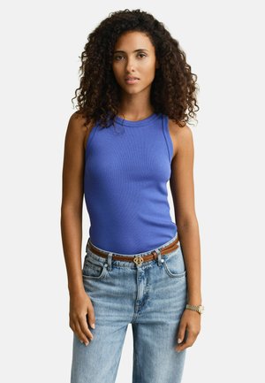 Vrouw met krullend haar, gekleed in een mouwloos blauw topje, lichtblauwe spijkerbroek, bruine riem en een horloge, staand tegen een witte achtergrond.