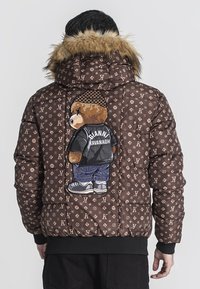Giubbotto imbottito marrone con cappuccio bordato di pelliccia, caratterizzato da un ricamo di un orsetto sulla schiena che indossa un giubbotto nero e jeans. Loghi sparsi.