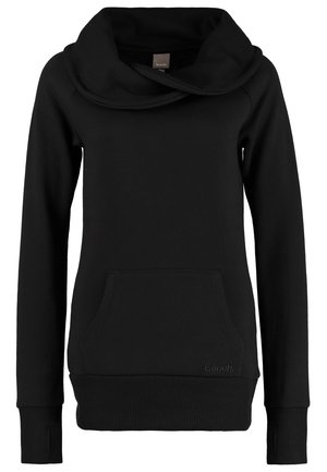 Person trägt einen schwarzen Sweatshirt mit Rollkragen und Daumenlöchern, ausgewaschene blaue Jeans und dunkelblaue Sneaker mit weißen Schnürsenkeln, steht vor einem schlichten Hintergrund.