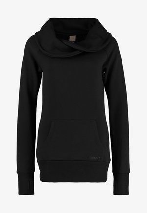 Person trägt einen schwarzen Sweatshirt mit Rollkragen und Daumenlöchern, ausgewaschene blaue Jeans und dunkelblaue Sneaker mit weißen Schnürsenkeln, steht vor einem schlichten Hintergrund.