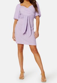 Robe courte violette en tissu texturé, avec des manches bouffantes, un décolleté en cœur et un détail à nouer à la taille. Présente un motif à carreaux.