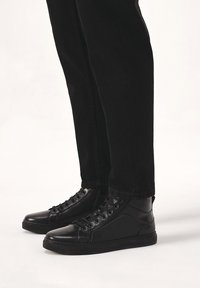 Črne visoke teniske iz gladke usnje, z okroglim prstom, s vezalkami in teksturiranimi stranskimi ploščami. V kombinaciji s črnimi kavbojkami.