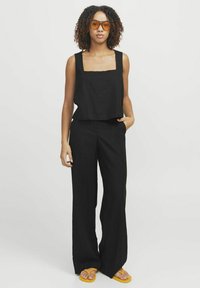 Zwarte linnen crop top met vierkante halslijn gecombineerd met brede zwarte pantalons. Model draagt oranje zonnebril en gele sandalen.