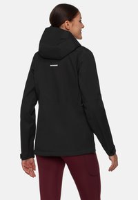 Donna di spalle che indossa una giacca a vento nera con cappuccio e logo "Mammut" e pantaloni attillati borgogna, su uno sfondo leggero e uniforme.