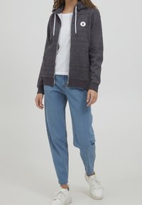 Joven con una sudadera con cremallera gris oscuro, camiseta blanca, jeans azul claro y zapatillas blancas, de pie contra un fondo liso.