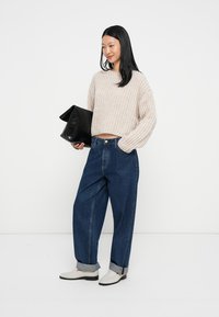 Maglione cropped beige in maglia, jeans blu a vita alta risvoltati alla caviglia, pochette nera e stivaletti bianchi alla caviglia. Outfit semplice e casual.