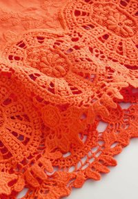 Nahaufnahme von leuchtend orangefarbenem Stoff mit filigranen, floralen Häkelspitzenmustern und strukturierter Stickerei.