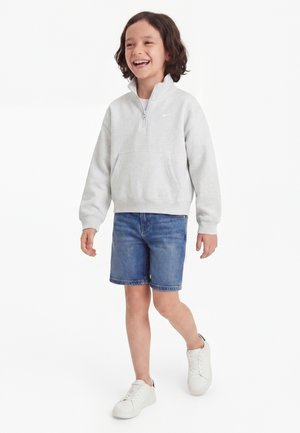 Enfant souriant et marchant, portant un sweat-shirt gris clair à zip quart, un short en denim et des baskets blanches sur fond blanc.