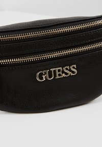 Sort lædertalje taske med flere lynlåslommer og et markant sølv GUESS logo på fronten. Struktureret overflade.