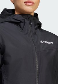 Čierna bunda Terrex so zipsom, kapucňou a štruktúrovanou tkaninou; na hrudníku sa nachádza biele logo.