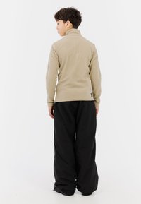 Beige fleece pullover met hoge kraag, gecombineerd met losse zwarte waterdichte broek. Structuurstof met ribbelmanchetten en -zoom.