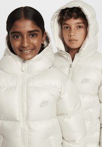 Chaquetas tipo puff en color crema, con un diseño acolchado, cuellos altos y logotipos de Nike. Tejido suave y brillante con capucha.