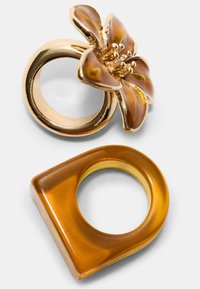 Goldfarbener Ring mit floralem Design in bernsteinfarbenen Tönen, neben einem quadratisch geformten Bernsteinring mit glatter Oberfläche und mittigem Durchbruch.