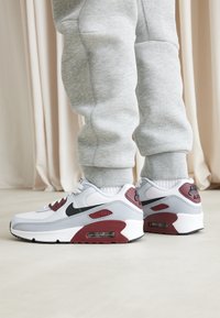Sapatilhas Nike Air Max 90 em branco, cinza e borgonha. Apresentam um swoosh preto, materiais de malha e couro, com amortecimento de ar visível.