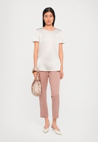 Una donna si trova di fronte a uno sfondo bianco, indossando una maglietta in satin beige a maniche corte, pantaloni cropped rosa polveroso, tacchi bianchi e tiene in mano una borsa in satin beige.