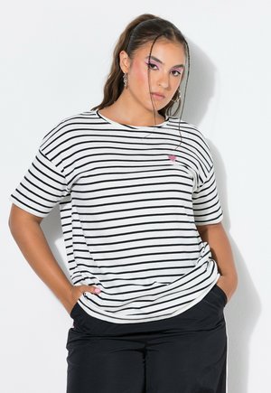 Junge Frau mit gestreiftem weiß-schwarzem T-Shirt mit kleinem pinkfarbenem Herzdetail, schwarzen Hosen und Händen in den Taschen vor weißem Hintergrund.